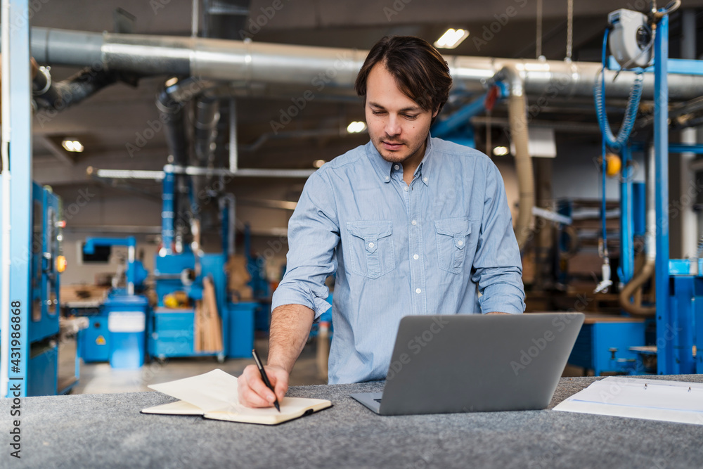 Male expertise with laptop writing in book while standing at industry Male expertise with laptop writing in book while standing at industry