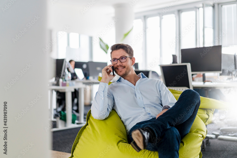 Handsome businessman talking through smart phone while sitting on bean bag at office Handsome businessman talking through smart phone while sitting on bean bag at office