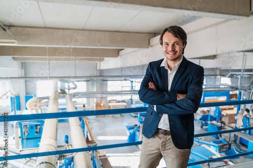 Young businessman with arms crossed smiling while standing at industry photo