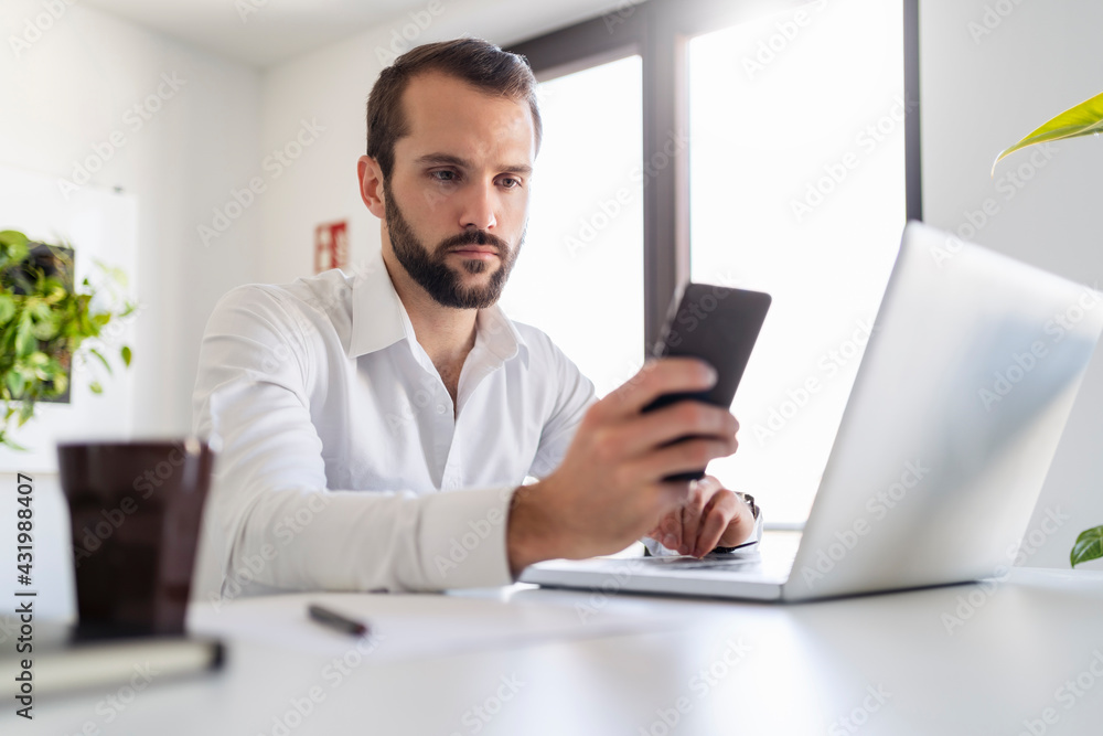 Young entrepreneur with laptop using mobile phone while sitting at office Young entrepreneur with laptop using mobile phone while sitting at office