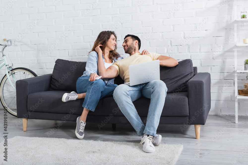 cheerful muslim man and happy woman looking at each other near laptop in living room. cheerful muslim man and happy woman looking at each other near laptop in living room.