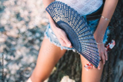 Horizontal close up image on woman legs in blue jeans shorts standing outdoors and holding a blue hand fan.