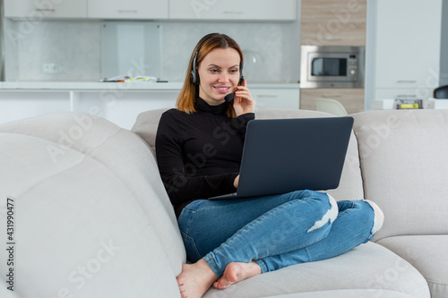 Young pretty girl works behind a laptop talking on the headset, sitting at home on the couch. High quality photo