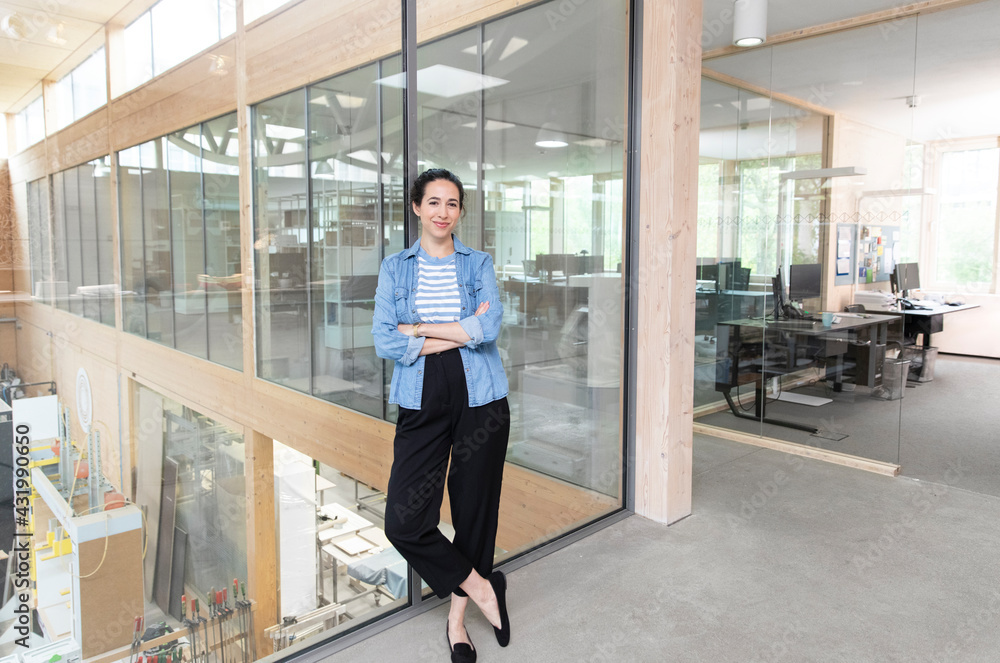 Confident female entrepreneur with arms crossed leaning on glass wall in factory Confident female entrepreneur with arms crossed leaning on glass wall in factory