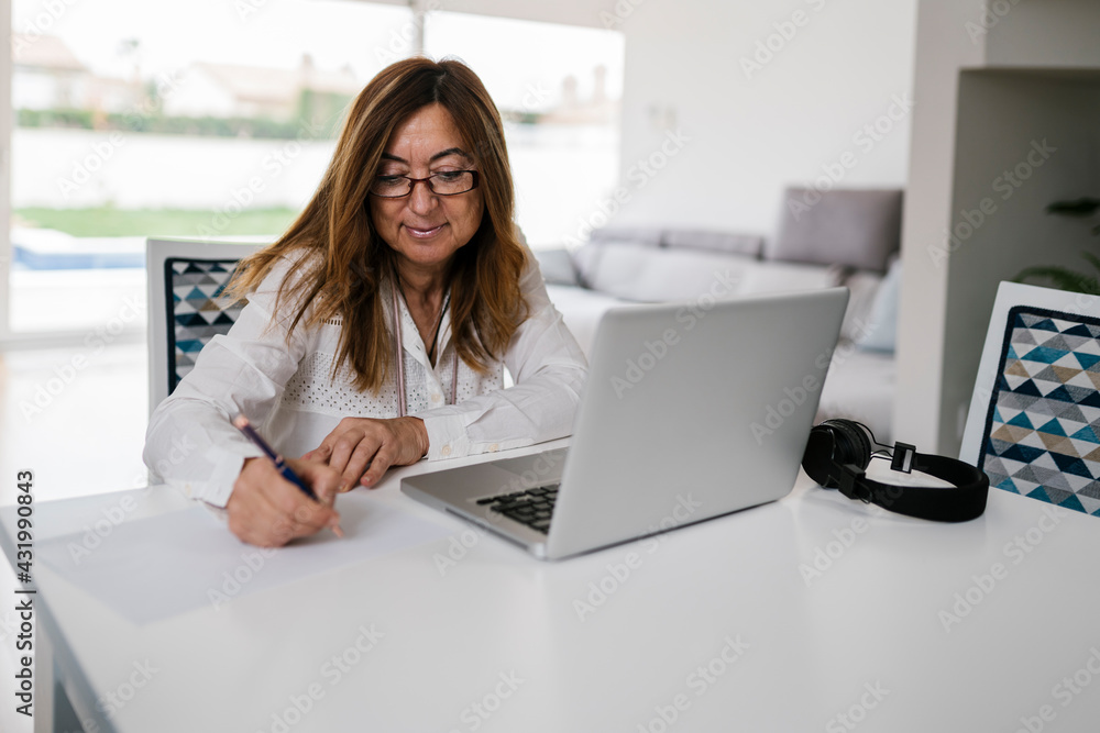 Female professional writing on paper while sitting with laptop and headphones at home Female professional writing on paper while sitting with laptop and headphones at home
