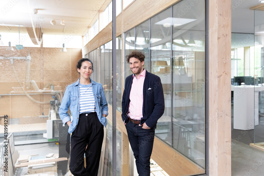 Male and female entrepreneurs with hands in pockets standing by glass wall in factory Male and female entrepreneurs with hands in pockets standing by glass wall in factory