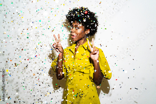 Woman showing peace sign and sticking out tongue while standing amidst confetti against white background photo