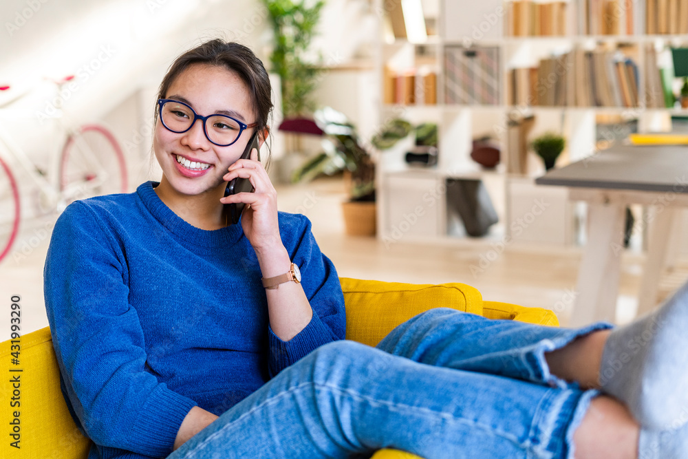 Smiling young woman talking on smart phone while sitting on chair at home Smiling young woman talking on smart phone while sitting on chair at home