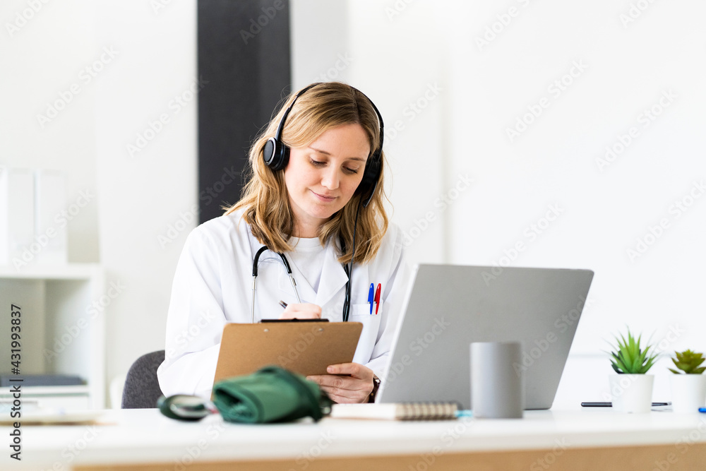 Female medical worker writing on clipboard while giving online consultation through laptop in medical clinic Female medical worker writing on clipboard while giving online consultation through laptop in medical clinic
