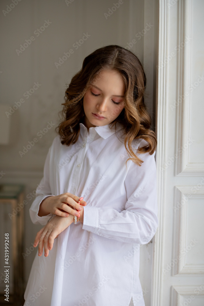 A beautiful little girl in a white shirt with long curly brown hair and blue eyes stands at the window and picks up a piece of jewelry A beautiful little girl in a white shirt with long curly brown hair and blue eyes stands at the window and picks up a piece of jewelry