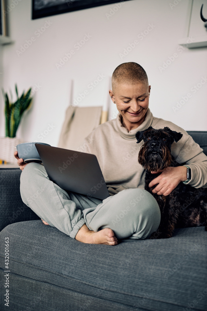 Smiling trendy senior woman with short hair sitting on the sofa with her dog and using technologies.