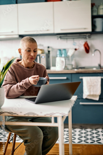 Attractive senior woman with short hair sitting at dining table in her kitchen, having healthy breakfast and using technologies.