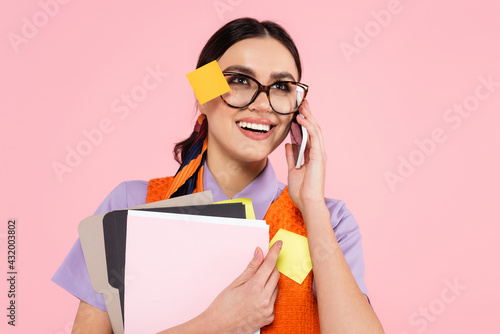 cheerful businesswoman talking on smartphone while holding documents isolated on pink.