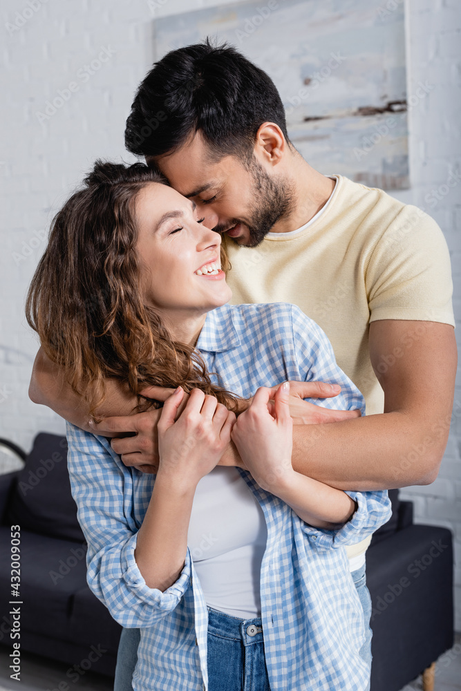 smiling multiethnic man and woman with closed eyes hugging at home.