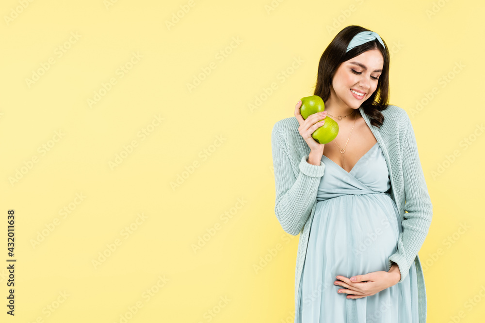 joyful pregnant woman holding fresh apples while touching tummy isolated on yellow. joyful pregnant woman holding fresh apples while touching tummy isolated on yellow.