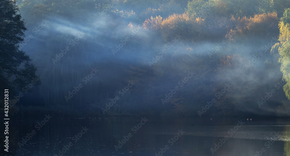 The sun shines through the fog on the forest around the lake.