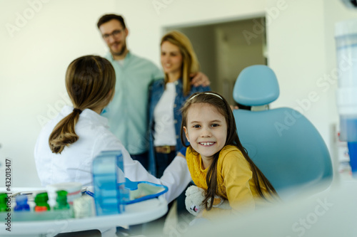 Smiling cute girl sitting in dental chair and looking at camera while parents talking with dentist.
