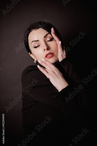 sexy brunette in black on a black background gracefully touches her face with her fingers