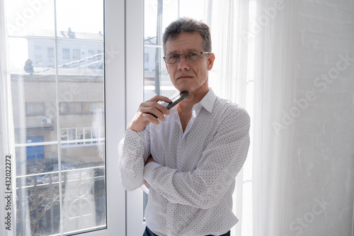 A man businessman in a white shirt and glasses with a phone stands at the window