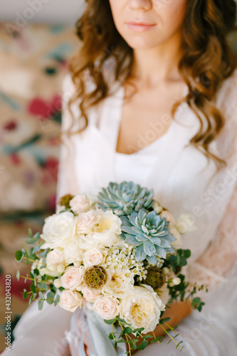 A bride in a lace robe sits on a sofa in a hotel room and holds a wedding bouquet in her hands, close-up