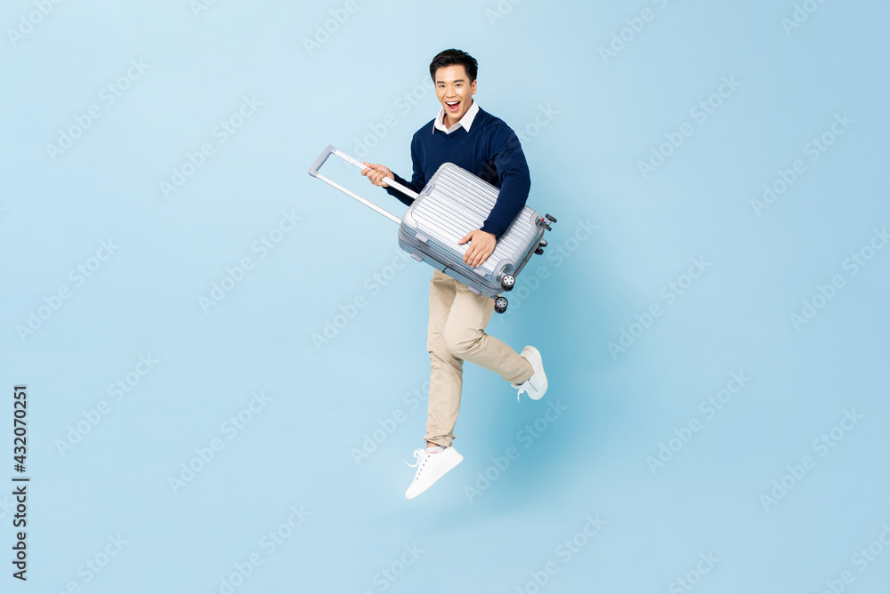 Jumping portrait of young fit healthy smiling handsome Asian tourist man with baggage ready to fly isolated on light blue studio background