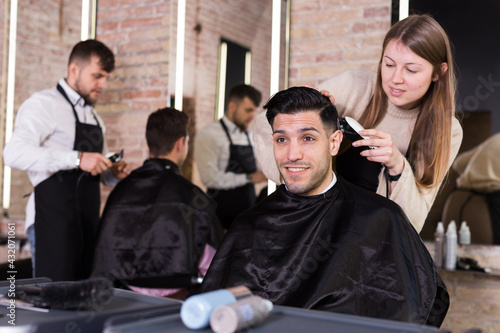 Female hairdresser cutting hair of male client with electric clipper in hair salon