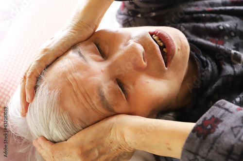 介護ベッドに横たわって目をつぶって両手で頭を抱えるシニア女性 photo