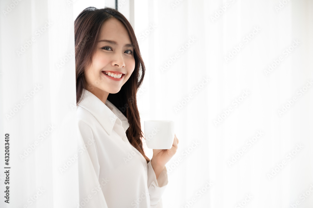 Young asian beautiful woman clothing white pyjamas and drink coffee beside window at morning in bedroom. She feeling happy fresh and smile relax Young asian beautiful woman clothing white pyjamas and drink coffee beside window at morning in bedroom. She feeling happy fresh and smile relax