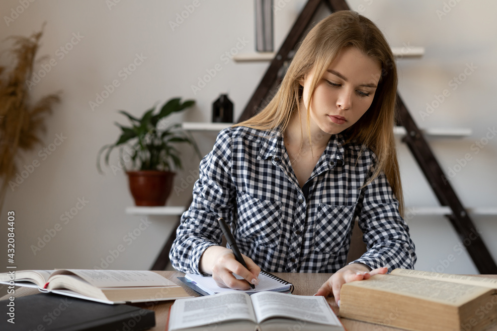 Motivated student sits with textbooks at a desk studying at home. A girl writes homework on mathematics in a notebook or a teacher prepares for a lecture in college remotely online. Motivated student sits with textbooks at a desk studying at home. A girl writes homework on mathematics in a notebook or a teacher prepares for a lecture in college remotely online.