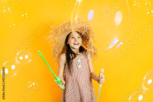 Little happy girl has fun blowing huge soap bubbles. Yellow bright wall background. Positive children's emotions.