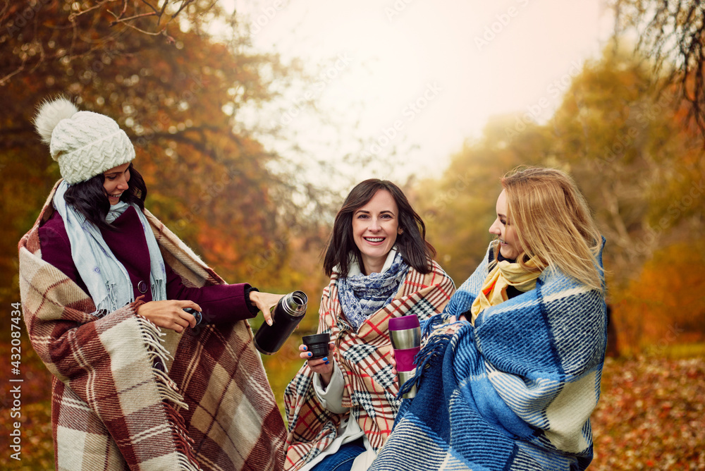 Three young cheerful girlfriends drinking hot tea in autumn forest