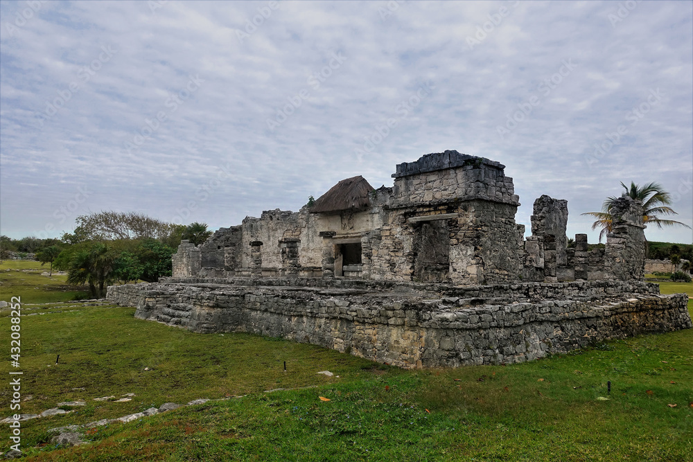 Ruins of the ancient Mayan city of Tulum. A dilapidated stone building against the backdrop of a cloudy sky. Around - green grass, tropical vegetation.
