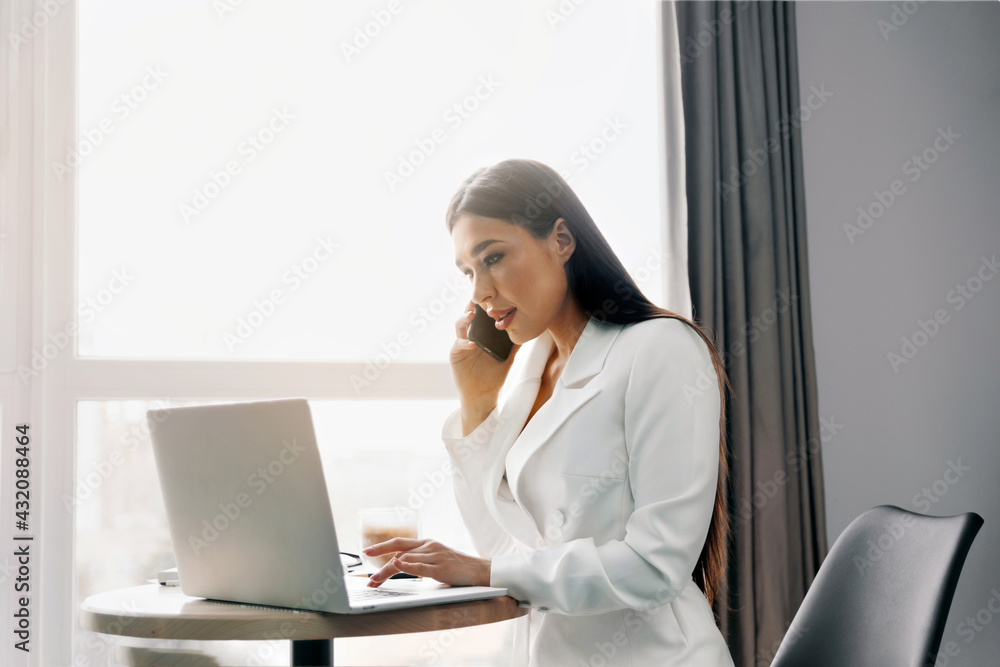 Beautiful woman talking on phone while working on laptop computer sitting at home managing her business via home office during Coronavirus or Covid-19 quarantine Beautiful woman talking on phone while working on laptop computer sitting at home managing her business via home office during Coronavirus or Covid-19 quarantine