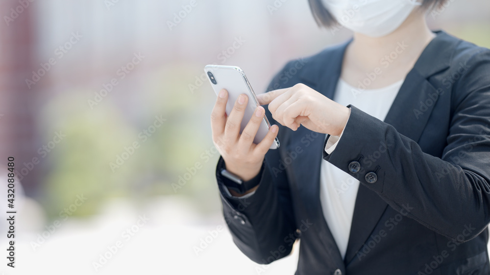 屋外でスマートフォンを操作するマスク姿のビジネスウーマン 屋外でスマートフォンを操作するマスク姿のビジネスウーマン