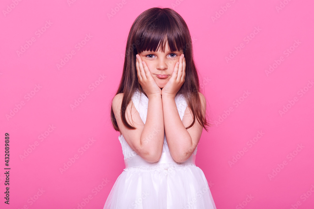 Adorable bored small girl looking at camera with tired expression, keeping hands on cheeks, does not know what to do, wearing beautiful white dress, isolated over pink background. Adorable bored small girl looking at camera with tired expression, keeping hands on cheeks, does not know what to do, wearing beautiful white dress, isolated over pink background.