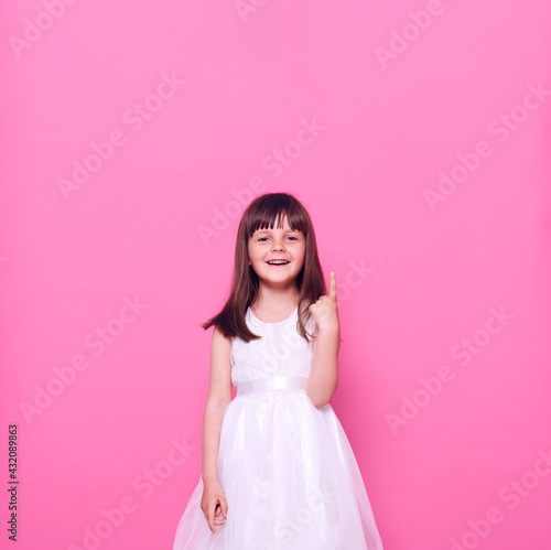 Attractive happy positive little kid wearing beautiful white dress pointing up with finger, looking at camera, copy space for promotion, isolated over pink background.