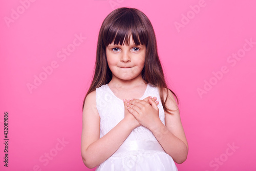 Charming honest little girl wearing white dress looking at camera, keeping hands on chest, has grateful expression, swearing, promising, isolated over pink background. photo