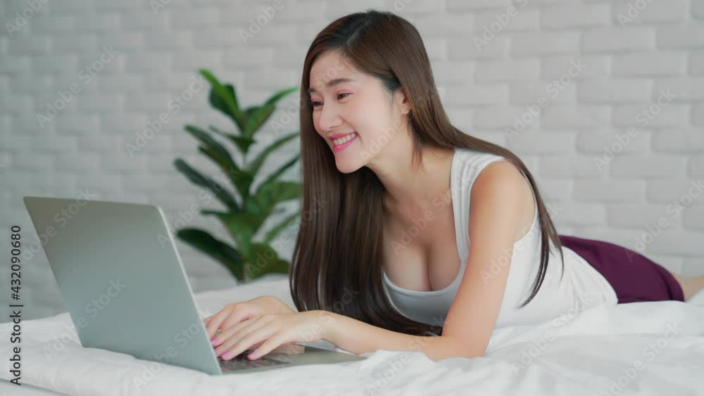 Smiling Asian woman resting in bed. Girl look at laptop screen enjoy pleasant personal conversation using video conference application.Technology help life easy remote communication from home concept Smiling Asian woman resting in bed. Girl look at laptop screen enjoy pleasant personal conversation using video conference application.Technology help life easy remote communication from home concept