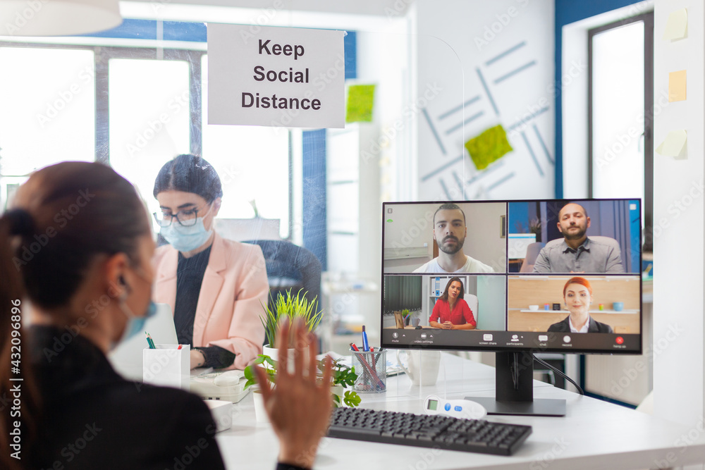 Business woman saying hello during remove video conference in new normal workplace wearing face mask as safety prevetion during global pandemic with covid10. Keeping social distancing from coworker. Business woman saying hello during remove video conference in new normal workplace wearing face mask as safety prevetion during global pandemic with covid10. Keeping social distancing from coworker.