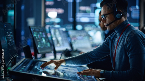 Portrait of Professional IT Technical Support Specialist Working on Computer in Monitoring Control Room with Digital Screens. Employee Wears Headphones with Mic and Talking on a Call.