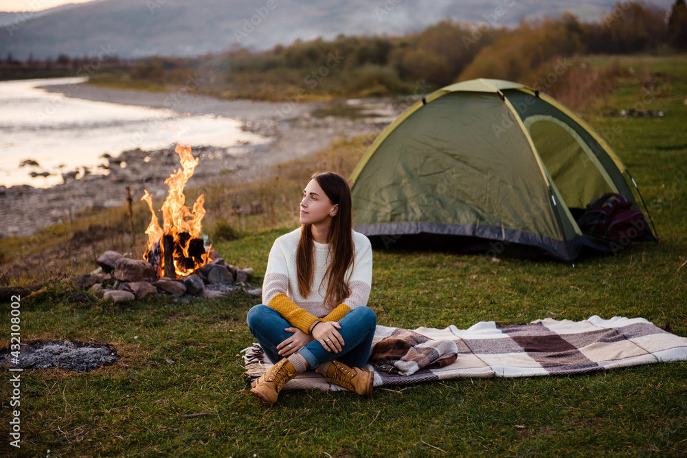 Front view of young brunette in the afternoon sits near a tent and enjoys mountain views. The campfire is lit on the side. Camping and hiking in the mountains.
