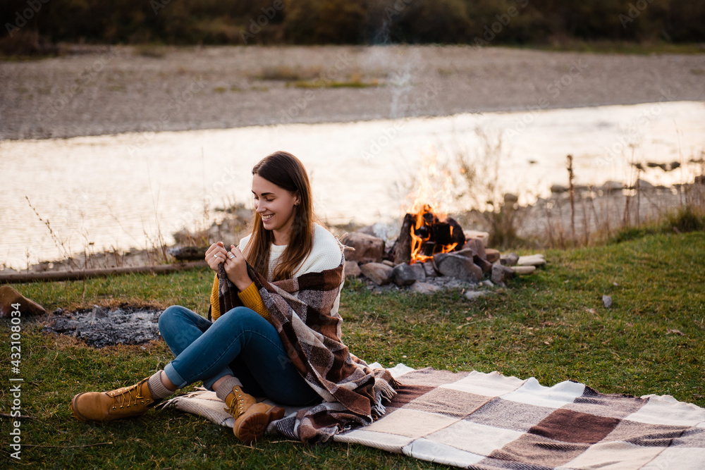 Young brunette tourist sitting on the grass. Woman covered herself with a blanket and enjoys the morning nature. Rest by the river. The campfire on the backgrount. Hiking concept.