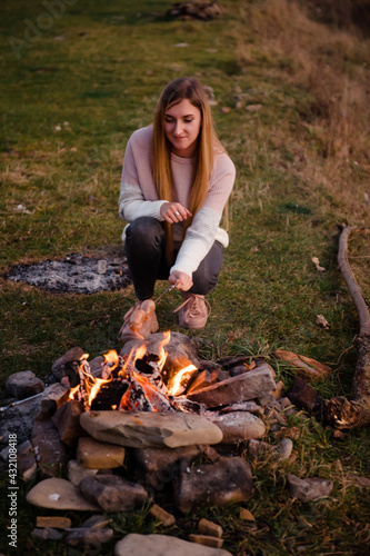 Camping in nature. Beautiful girl on a hike. A woman prepares sausages on a metal skewer on a fire. Green meadow and mountains on the background. Hiking and resting concept.