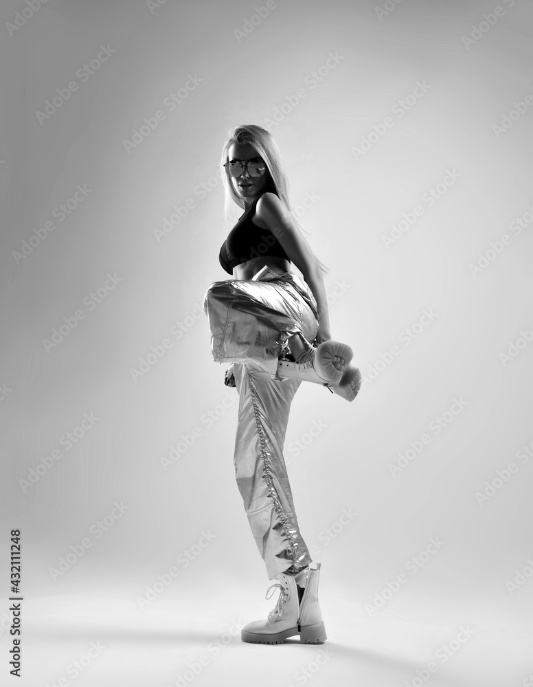 Obraz premium Black and white full-growth portrait of fitness trainer, sporty woman, hip-hop dancer in silver pants, black top and brutal shoes standing sideways kicking over background