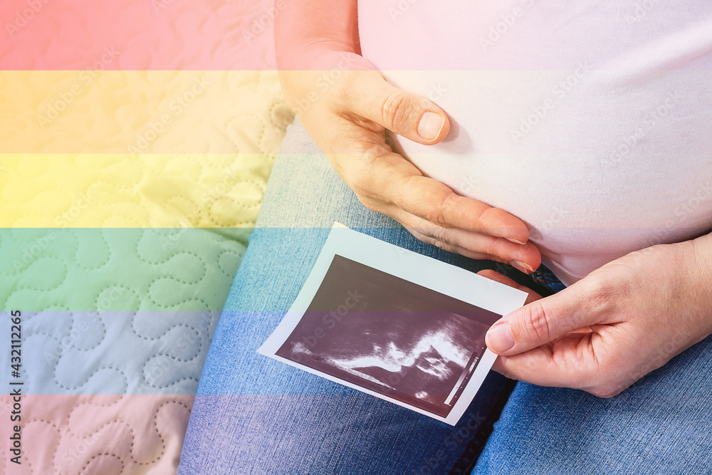 LGBT concept. Pregnant due to eco woman sitting on bed. One hand hugs her tummy and in other holds ultrasound photo of future baby. Lgbt flag colours covered photo. LGBT concept. Pregnant due to eco woman sitting on bed. One hand hugs her tummy and in other holds ultrasound photo of future baby. Lgbt flag colours covered photo.