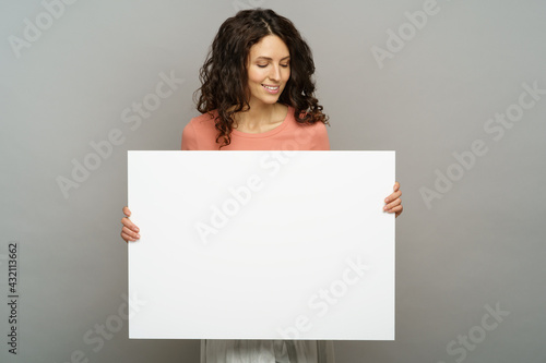 Excited young adult female hold blank placard looking down to empty copy space with pleasant smile. Beautiful millennial woman in pink outfit show while poster or banner isolated at studio background