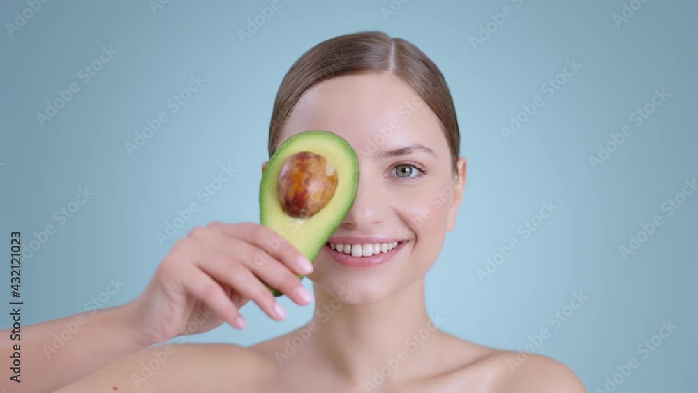 Portrait of millennial woman holding half of avocado in front of face and smilling while looking to camera . Female model with naked shoulders and light nude make up posing .Concept of skincare. Portrait of millennial woman holding half of avocado in front of face and smilling while looking to camera . Female model with naked shoulders and light nude make up posing .Concept of skincare.