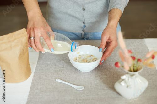 Female hand holding bottle pouring milk in cereal granola flakes bowl