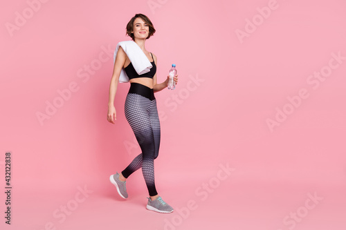 Full length photo of cheerful young fit woman walk hold water rest towel isolated on pink color background