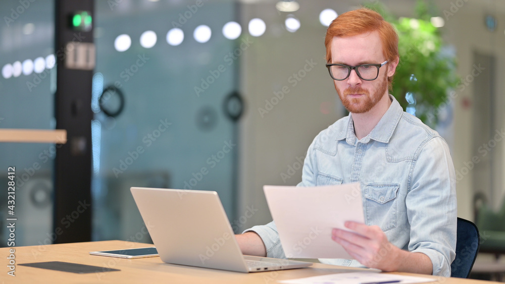 Young Redhead Man Working on Laptop with Documents in Office Young Redhead Man Working on Laptop with Documents in Office